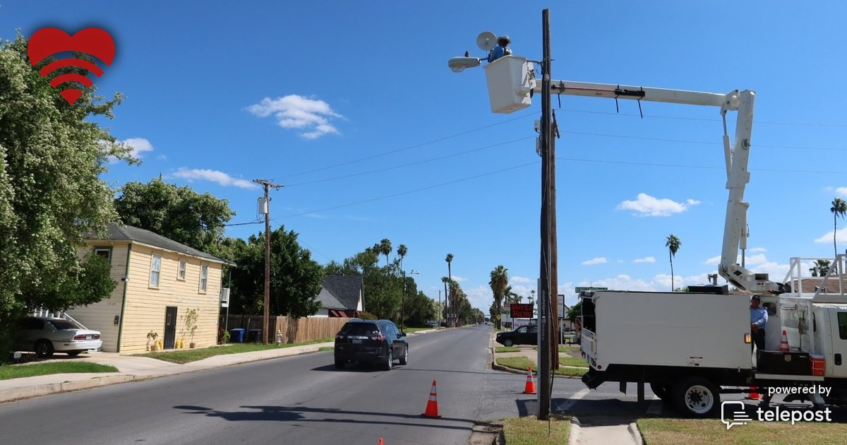 A small outdoor wireless access point mounted high on a city street light pole at dusk, with the empty street and sidewalk stretching away below it.