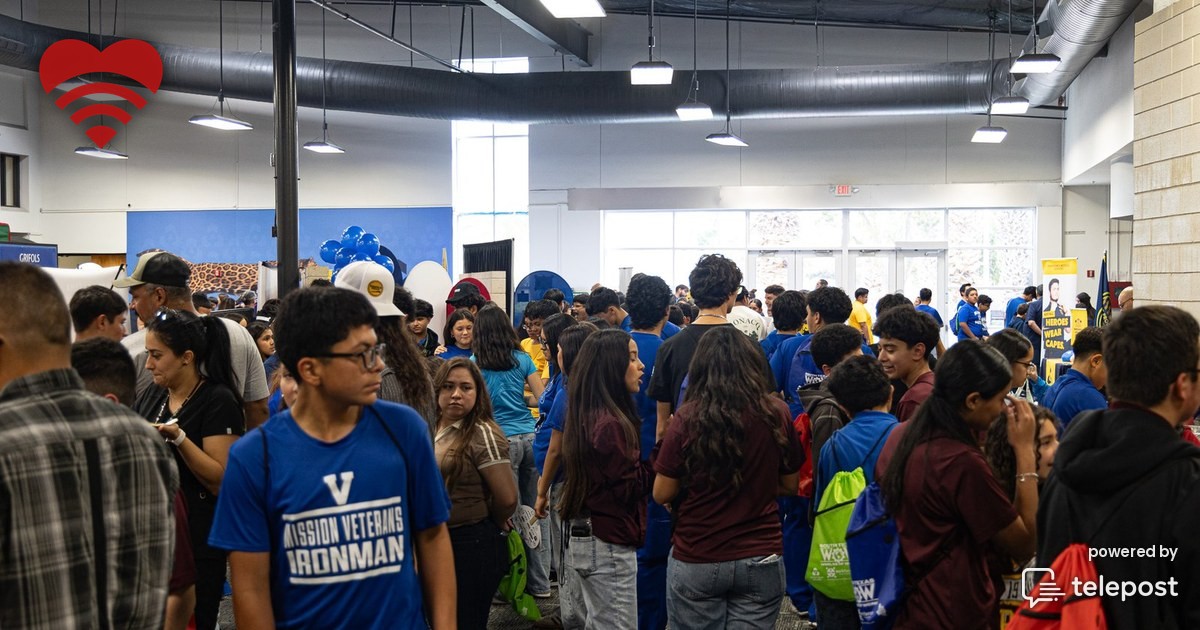 A folding table at a school job fair set with wireless access points, a spectrum analyzer and small handheld test tools, with a teenager leaning in to look at the gear while the gymnasium floor stretches away empty in the foreground.
