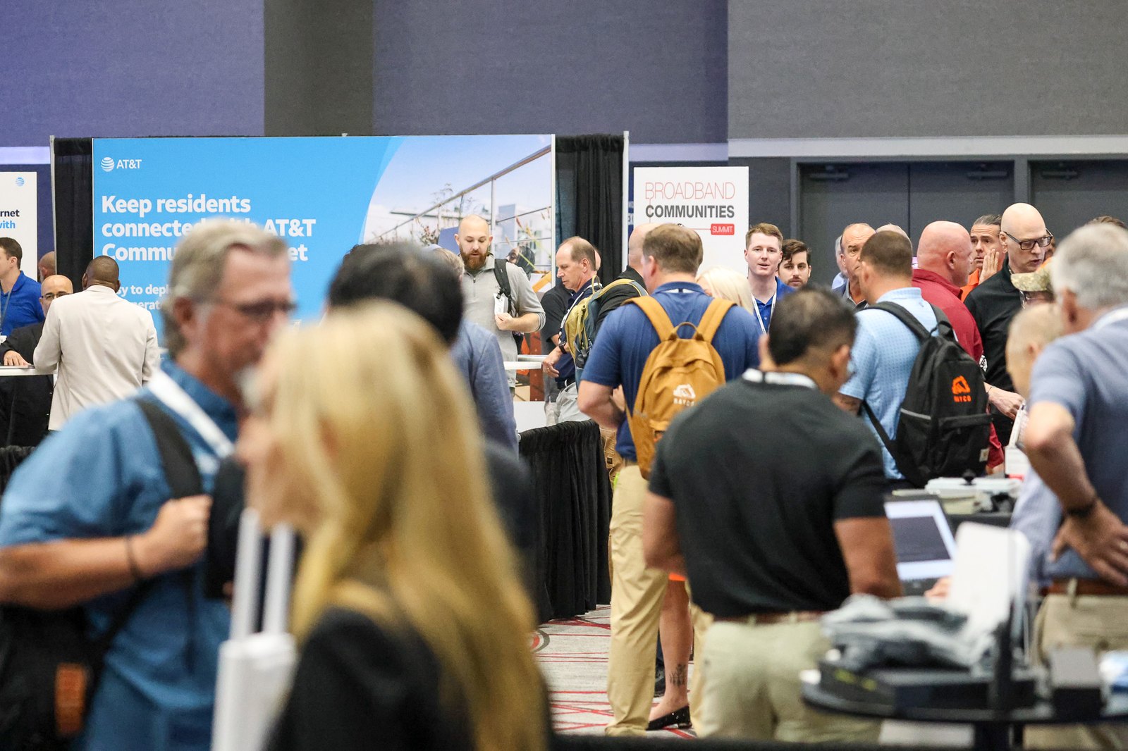 Crowd of conference attendees with backpacks and lanyards mingling on a trade show floor beside AT&T and Broadband Communities Summit banners.