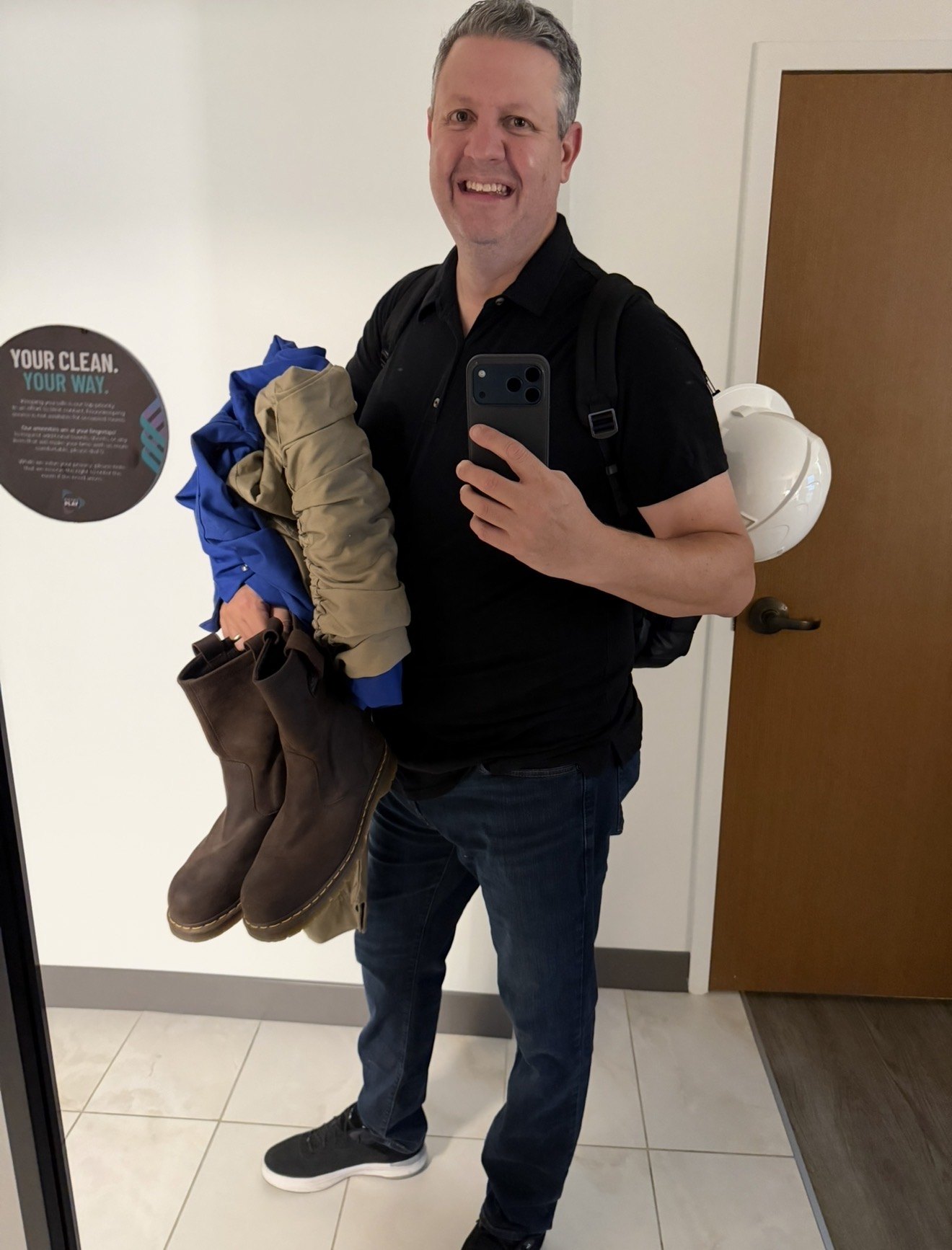 A smiling man in a black polo takes a mirror selfie while carrying brown boots, blue and tan jackets, a backpack and a white hard hat, beside a wooden door.