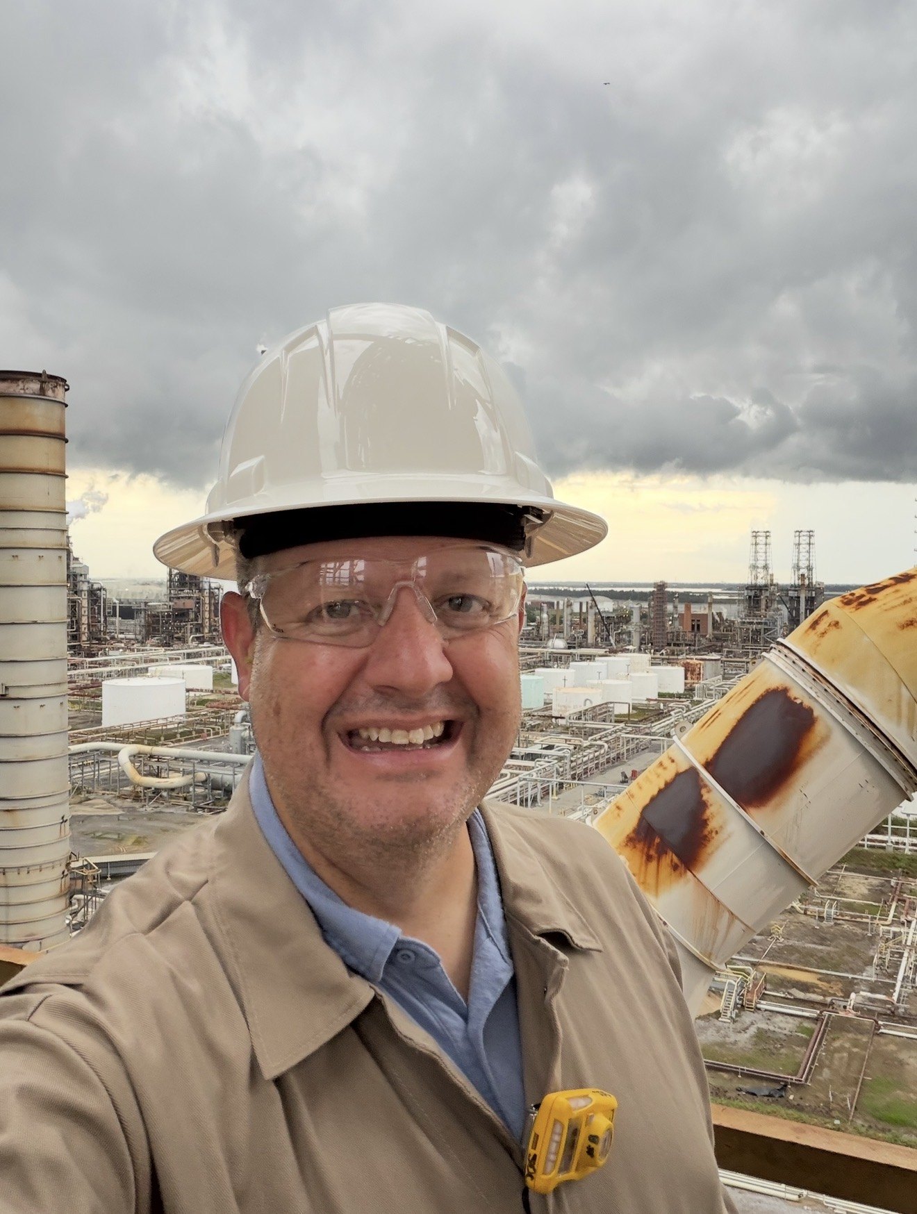 Smiling man in a white hard hat, safety glasses and tan coveralls takes a selfie high above a sprawling refinery, with a rusted duct and grey clouds behind.