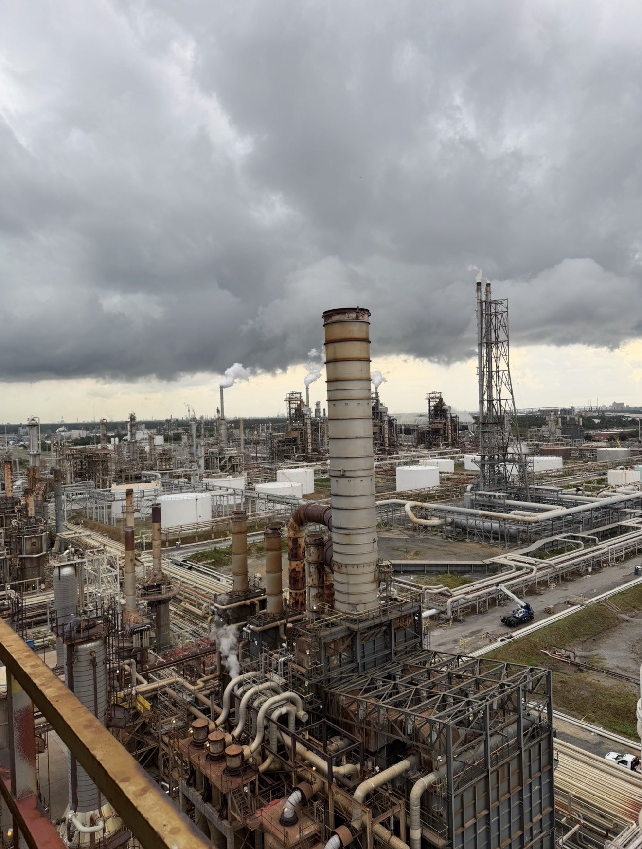 Sprawling oil refinery with tall rusted stacks, pipe racks and white storage tanks releasing steam plumes under heavy grey storm clouds, seen from a high walkway.
