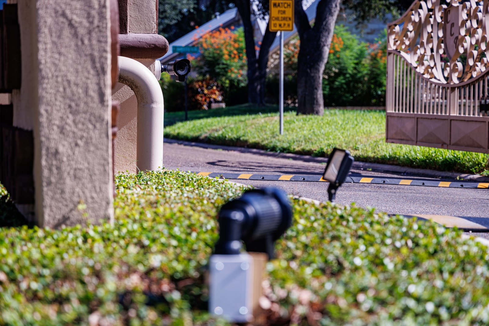 A small black security camera mounted on a white pipe against a stucco pillar, overlooking a driveway with a speed bump, hedges, an ornate metal gate, and a yellow "Watch for Children" sign.