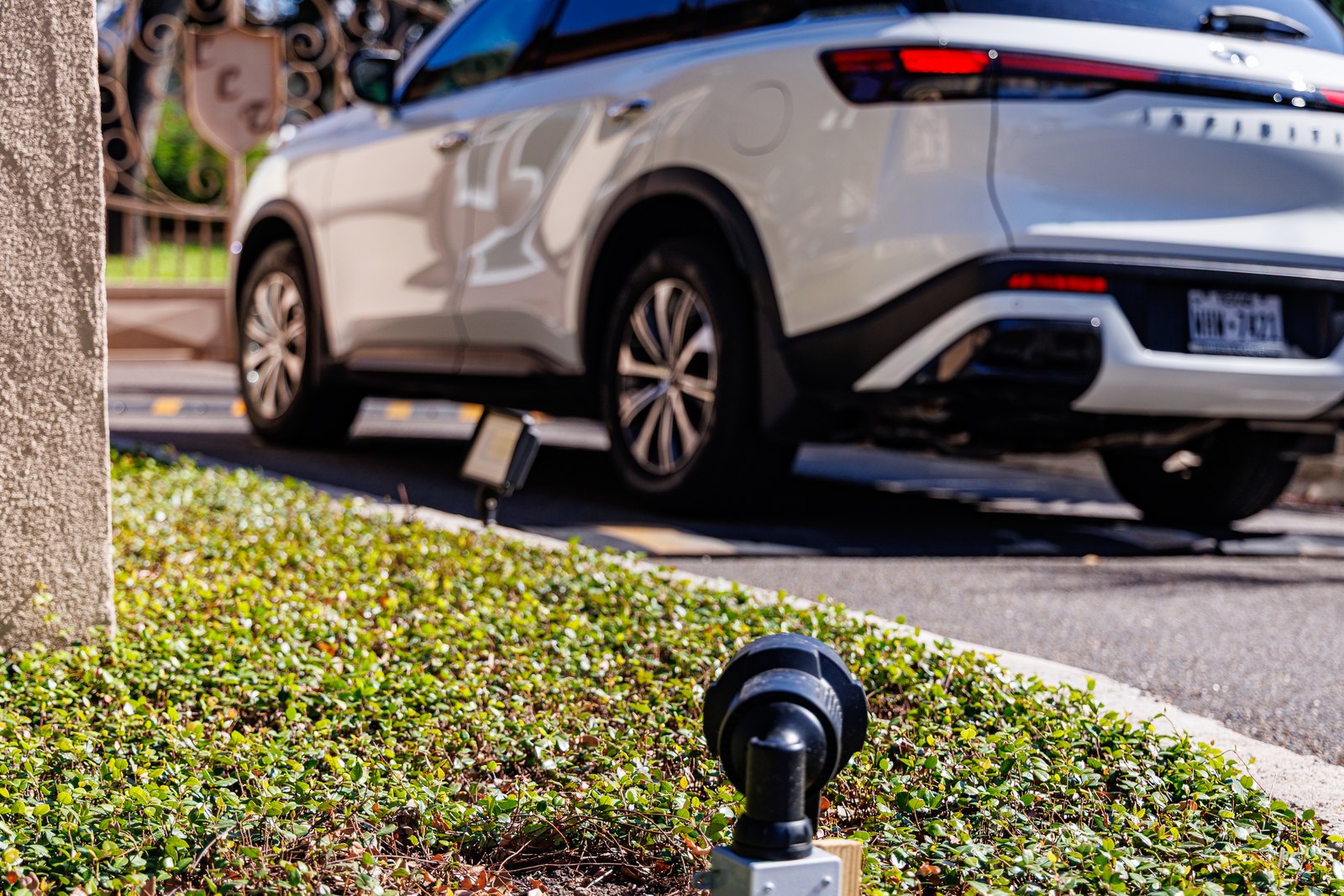 A black landscape light fixture sits in low green groundcover beside a stone pillar, with a white Infiniti SUV blurred on the pavement behind it.