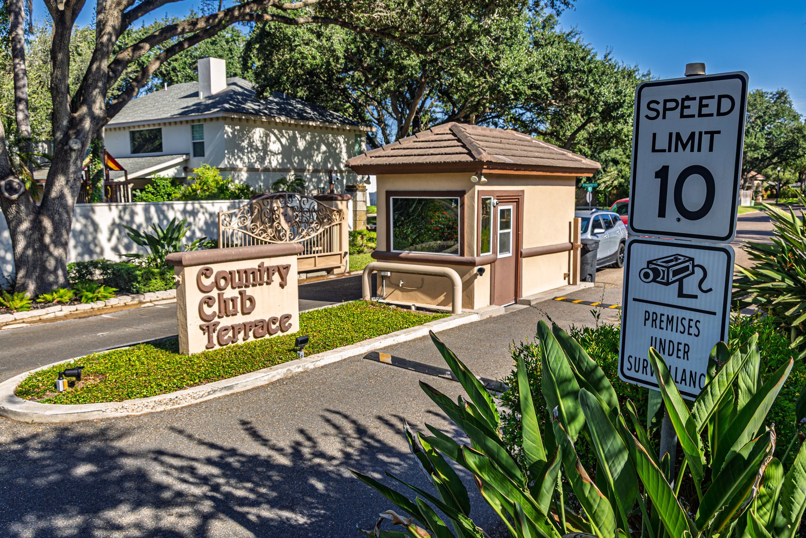 Gated community entrance with a stucco guard booth, "Country Club Terrace" stone sign, and a 10 mph speed limit and surveillance sign under shady oak trees.