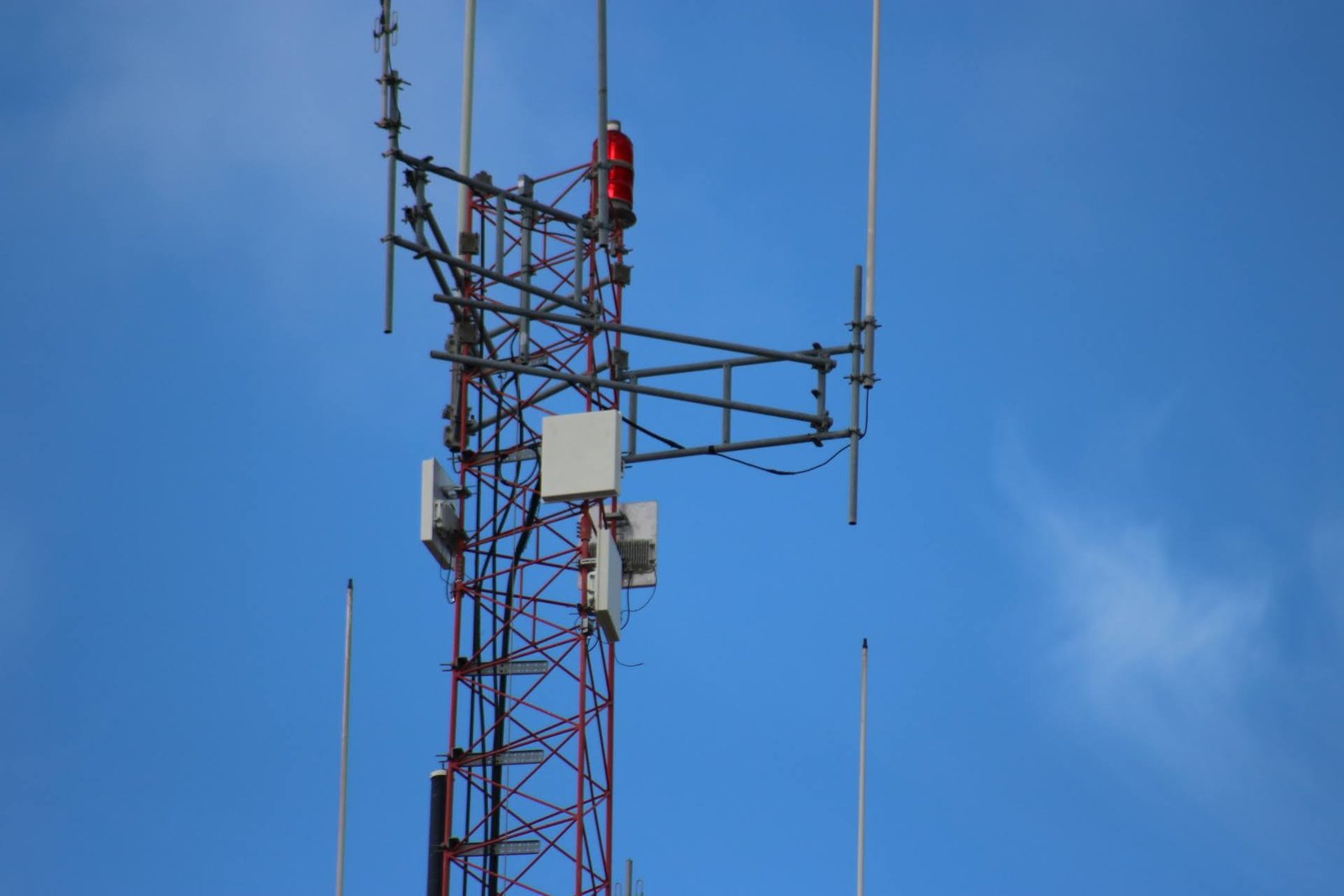 Red and white lattice telecommunications tower topped with a red beacon light, carrying panel antennas, mounting arms and slender whip antennas against a blue sky.