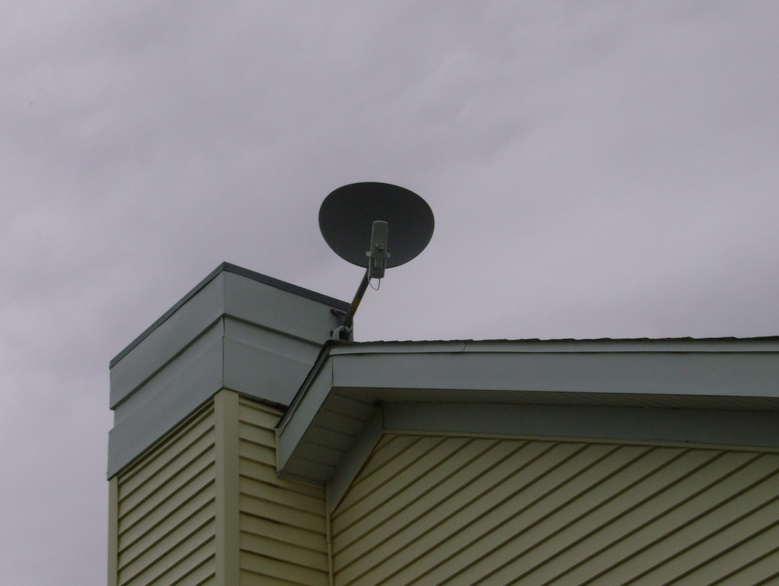 A small dark satellite dish mounted on a bracket at the corner roof edge of a house with cream vinyl siding, set against an overcast gray sky.