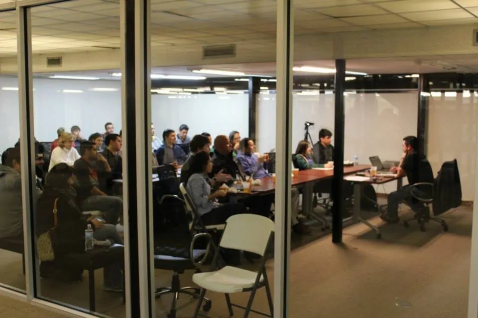 A group of about twenty people seated around tables in a glass-walled conference room, facing a person with a laptop at the front.