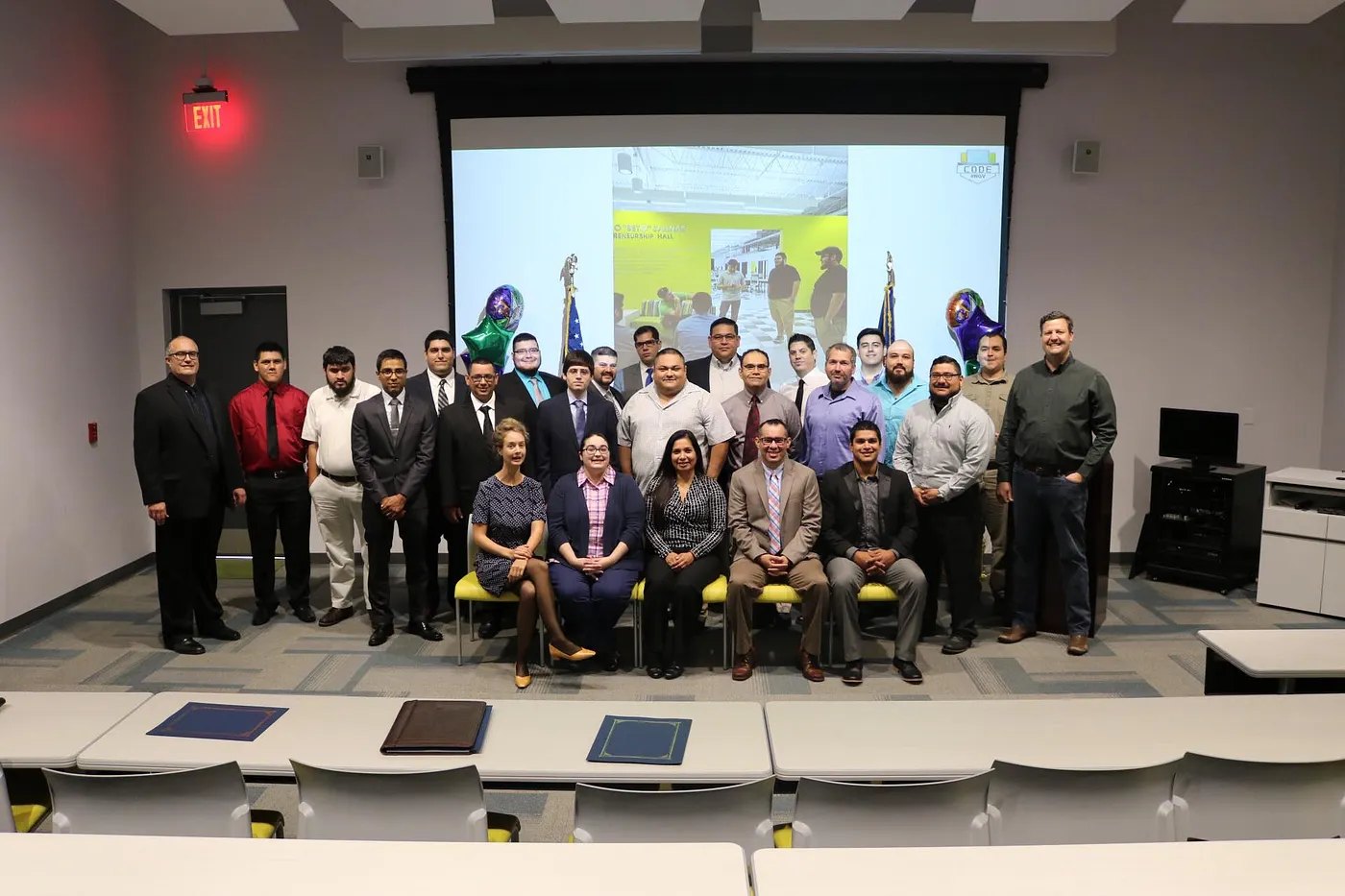 A group of about 25 people in business attire pose for a photo, five seated in front, in a lecture hall with a projection screen and balloons behind them.