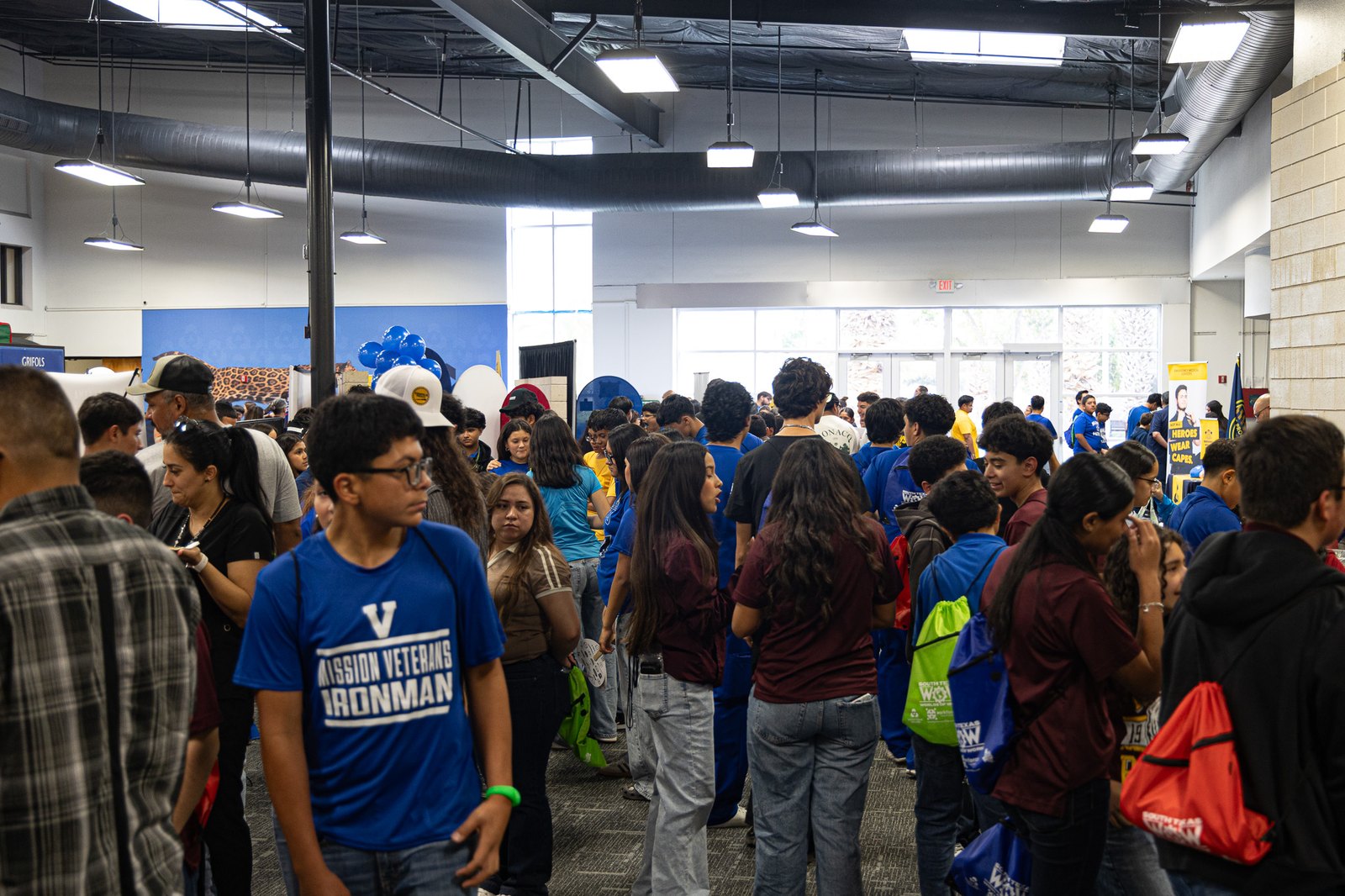 A large crowd of students, many in blue and maroon shirts with drawstring backpacks, mingles in a bright indoor hall with exposed ductwork and event banners.