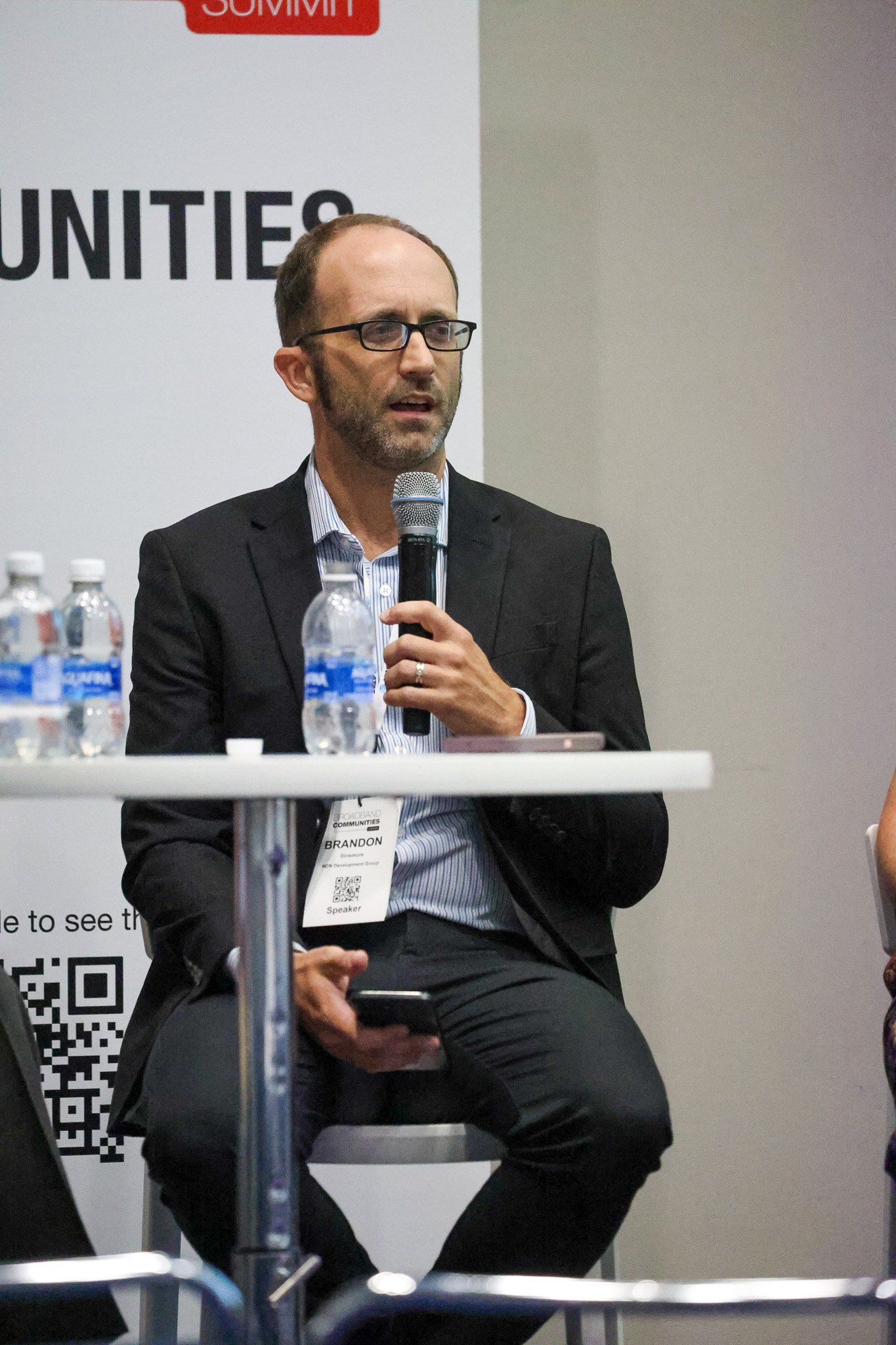A bearded man in glasses and a dark suit speaks into a handheld microphone while seated at a small white table with water bottles, in front of a Broadband Communities Summit banner.