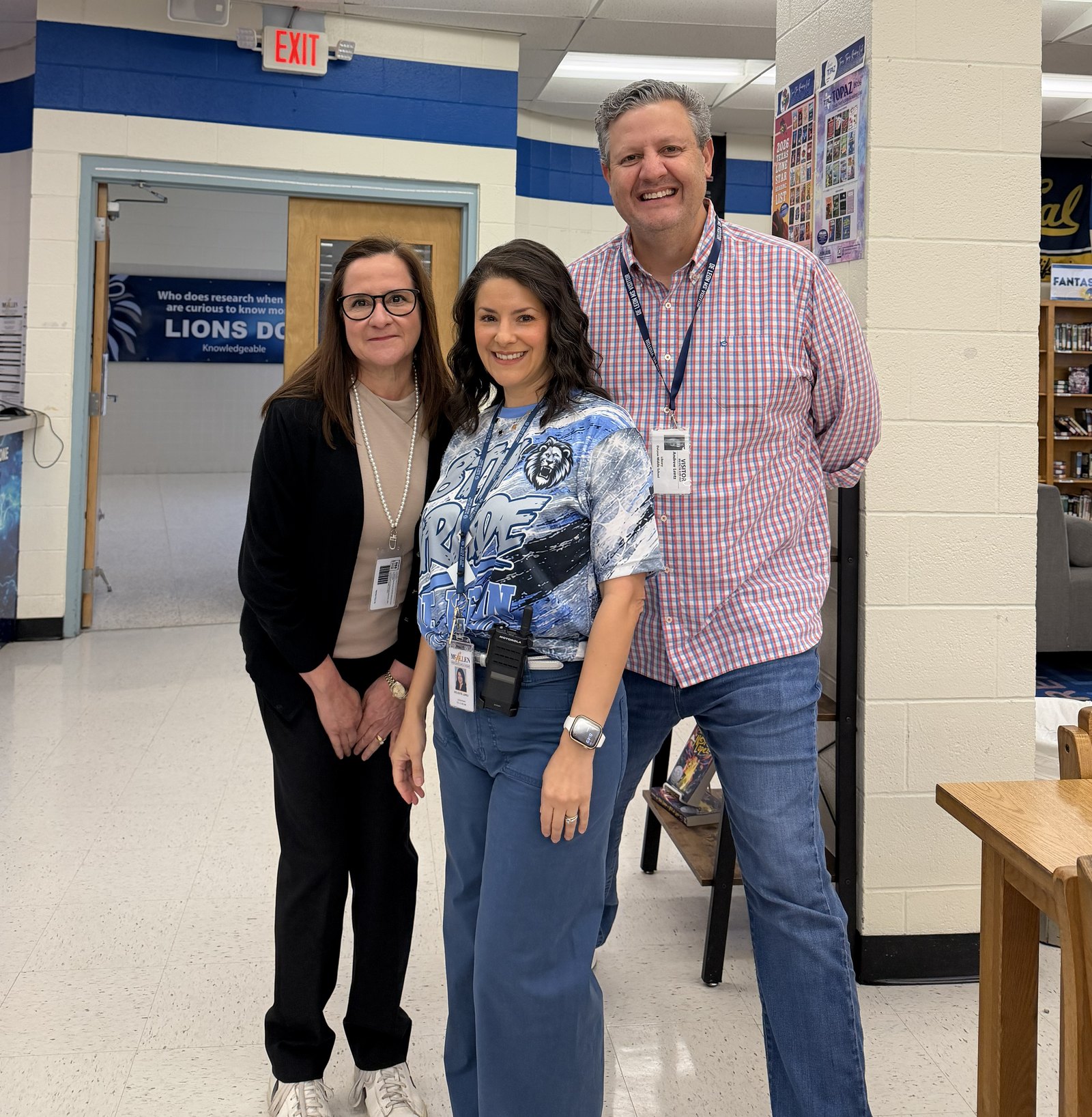 Three adults stand smiling side by side in a school library, two wearing staff lanyards and one in a blue Lions spirit shirt with a radio clipped on.