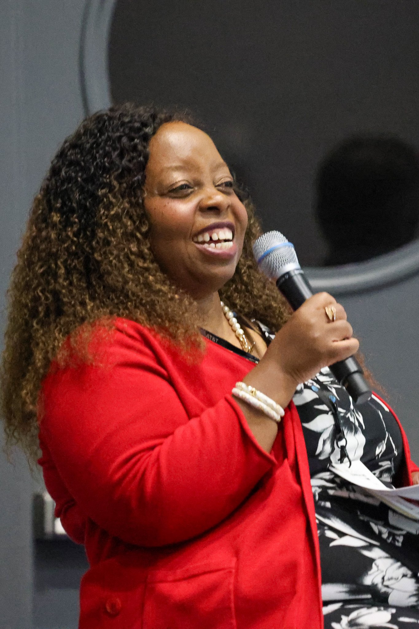 A smiling woman with long curly hair speaks into a handheld microphone, wearing a red blazer over a black-and-white floral dress, against a grey backdrop.