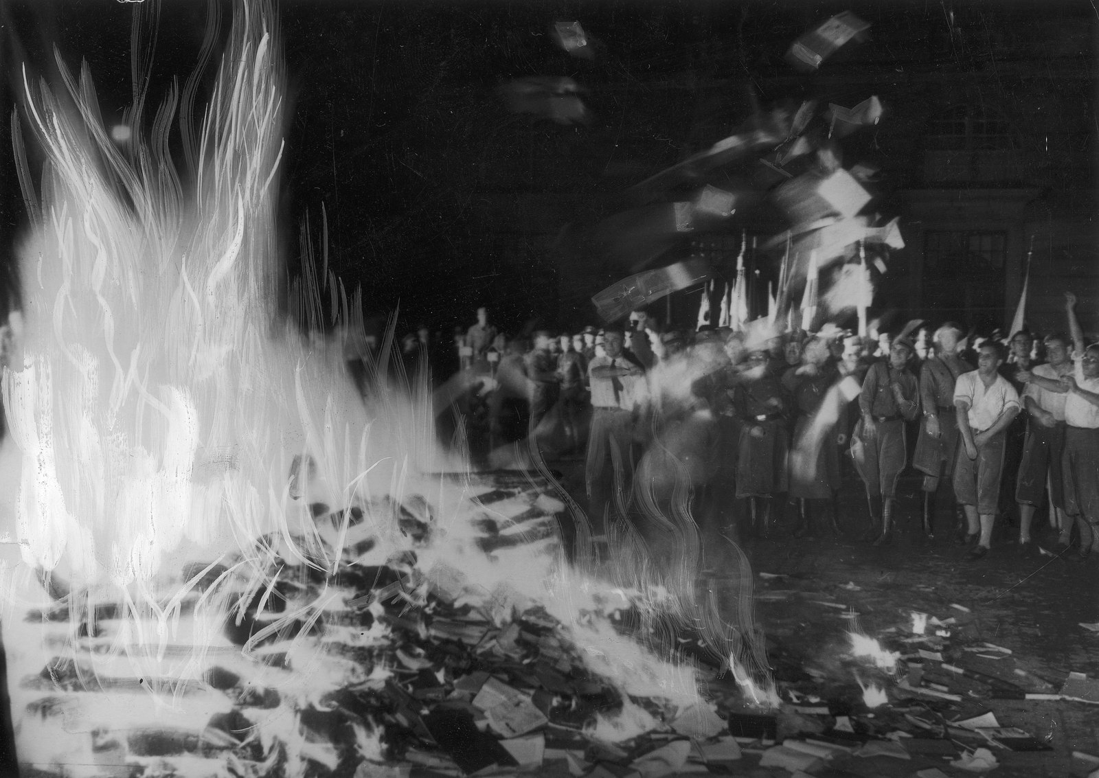 Black-and-white night photo of a large bonfire of books and papers, with a crowd of men in uniforms and shirts watching behind flags and thrown pages.