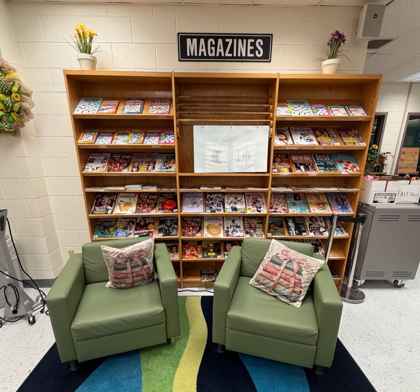 Two green armchairs with book-print pillows sit on a striped rug in front of a wooden shelving unit of magazines under a "MAGAZINES" sign.