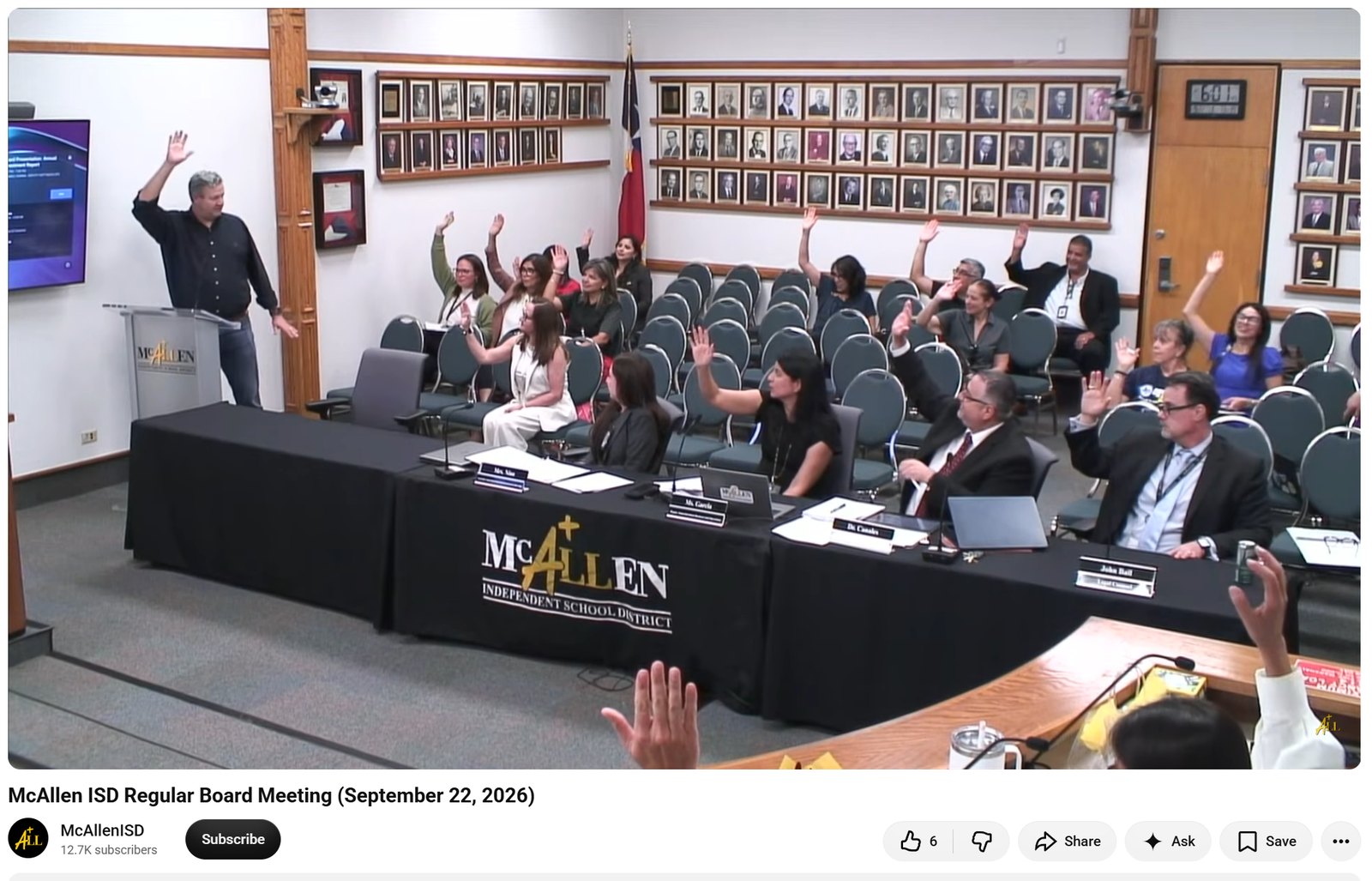 Video still of a man at a podium raising his hand while seated attendees raise theirs in a McAllen ISD board meeting room lined with framed portraits.