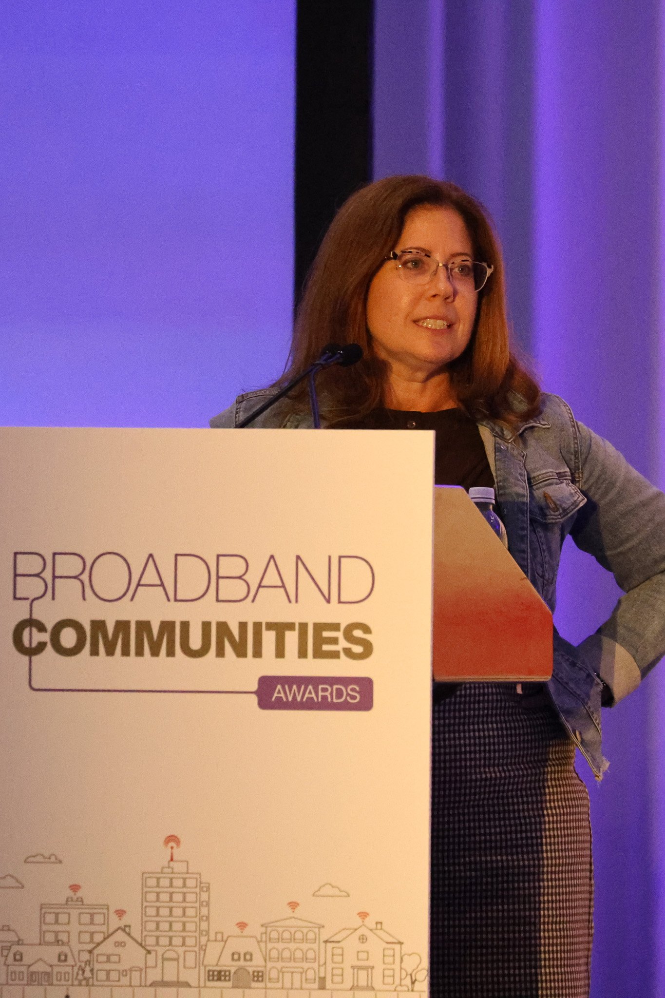 A woman with long brown hair and glasses speaks at a podium bearing the "Broadband Communities Awards" logo, in front of a purple-lit curtain.