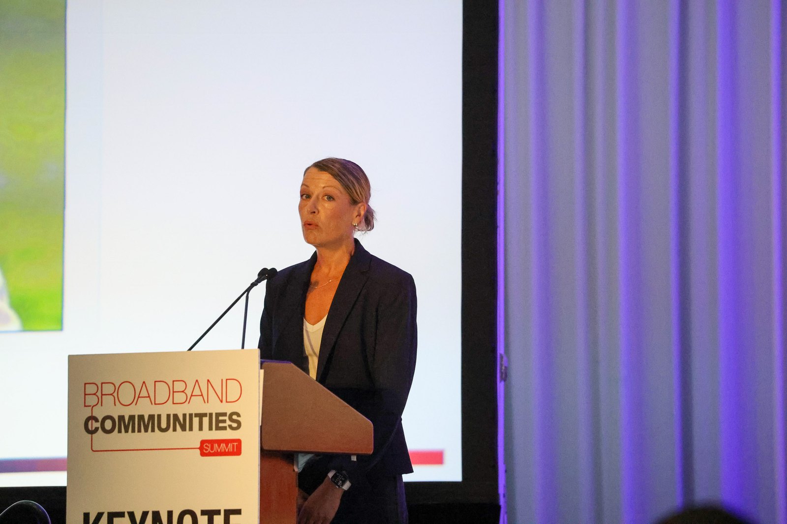 A woman in a dark blazer speaks into a microphone at a podium labeled "Broadband Communities Summit Keynote," with a projection screen and purple curtain behind.