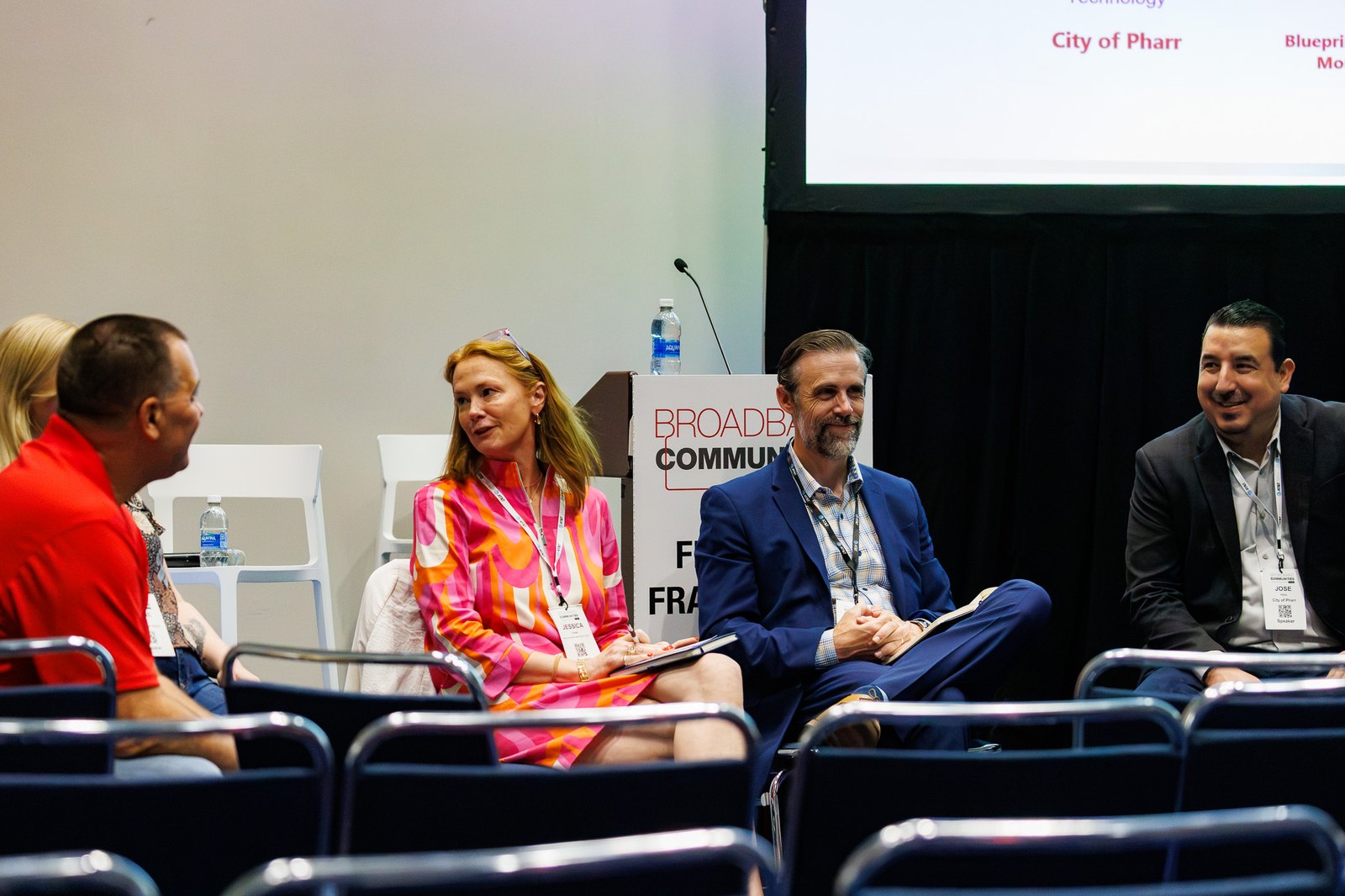 Four panelists seated on stage smiling and talking at a Broadband Communities conference, with a podium, microphone and presentation screen behind them.