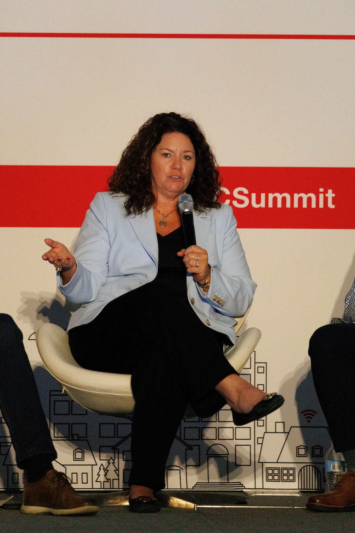 A woman with curly brown hair in a light blue blazer and black outfit speaks into a handheld microphone while gesturing, seated in a cream chair before a red-and-white "Summit" backdrop.
