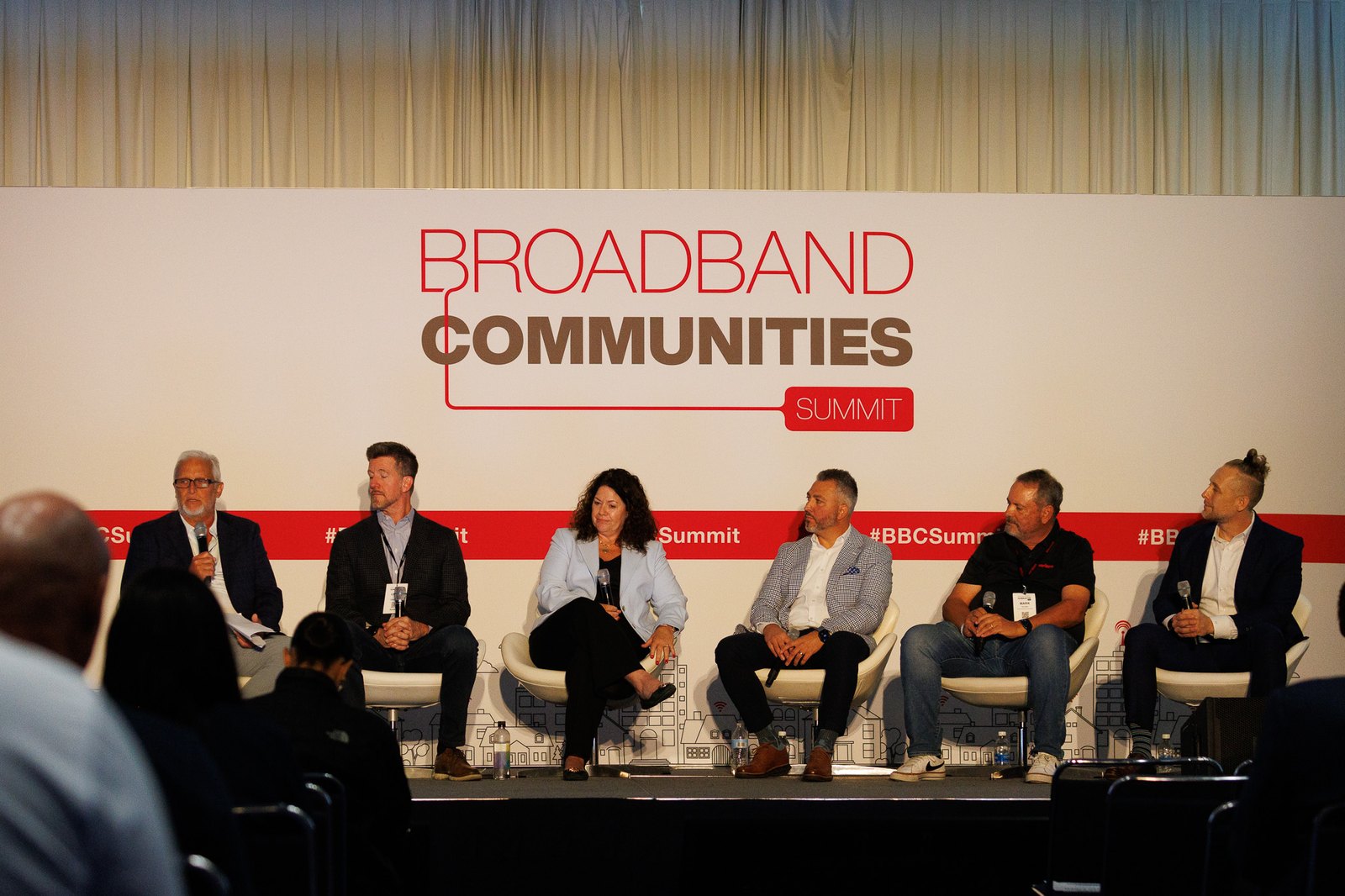 Six panelists seated in white chairs on stage, one speaking into a microphone, in front of a Broadband Communities Summit backdrop.